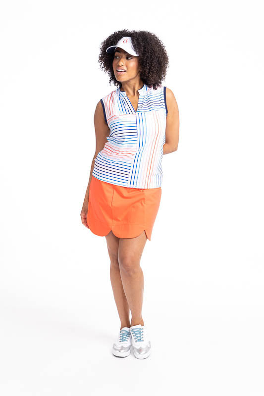 Full front view of a smiling woman wearing the Sandy Par Sleeveless Golf Top in Sun Stripe Print, the On The Edge Golf Skort in Coral Red, and the No Hat Hair Visor in White. The Sun Stripe print consists of a mix of horizontal and vertical stripes in cor