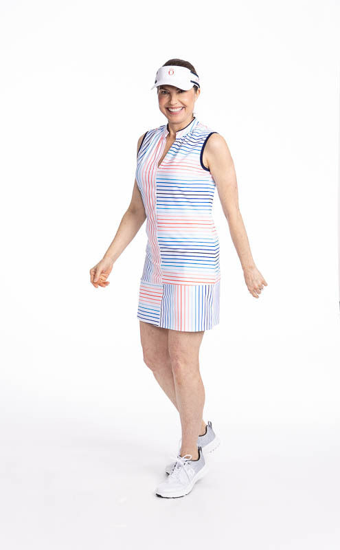 Full front view of a smiling woman wearing the Sandy Par Sleeveless Golf Dress in Sun Stripe print and the No Hat Hair Visor in White. The print consists of a mix of horizontal and vertical stripes in coral red, pacific blue, and navy blue on a white back