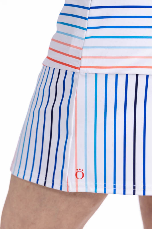 Close left side view of the hemline on the Sandy Par Sleeveless Golf Dress in Sun Stripe print. The print consists of a mix of horizontal and vertical stripes in coral red, pacific blue, and navy blue on a white background. The low-banded collar and armho