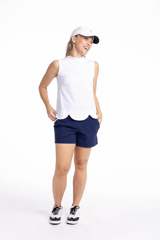 Full front view of a smiling woman wearing the On The Edge Sleeveless Golf Top in White, the Carry My Cargo Shorts in Navy Blue/White, and the We've Got You Covered Hat in White. This top features a back zipper and a feminine scallop design along the heml