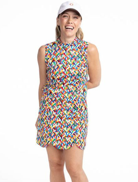 Front view of a smiling woman wearing the On The Edge Sleeveless Golf Dress in K All Day print. The K All Day print is an abstract confetti-style mirrored K-shaped pattern in red, orange, yellow, green, blue, black and white.