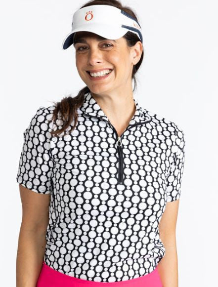 Close front view of a smiling woman wearing the No Hat Hair Visor in White and the Keep It Covered Short Sleeve Golf Top in Spiral Floral Print. This top is trimmed in black down the front quarter zipper. The print is a black and white retro-inspired abst