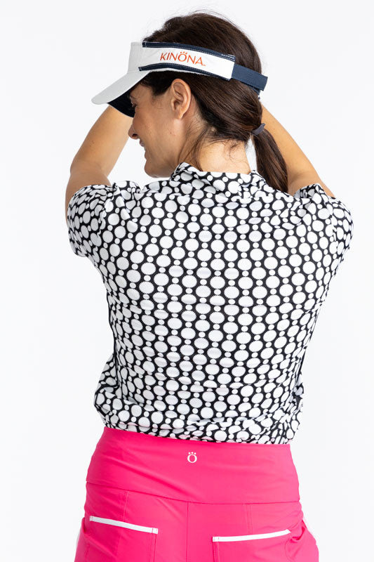 Close back view of a woman wearing the No Hat Hair Visor in White and the Keep It Covered Short Sleeve Golf Top in Spiral Floral Print. This top is trimmed in black down the front quarter zipper. The print is a black and white retro-inspired abstract flow