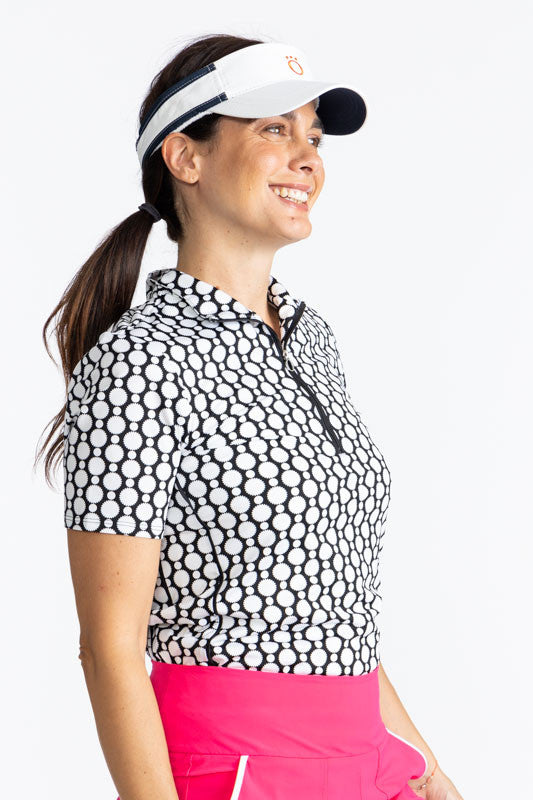 Right side view of a smiling woman wearing the No Hat Hair Visor in White and the Keep It Covered Short Sleeve Golf Top in Spiral Floral Print. This top is trimmed in black down the front quarter zipper. The print is a black and white retro-inspired abstr
