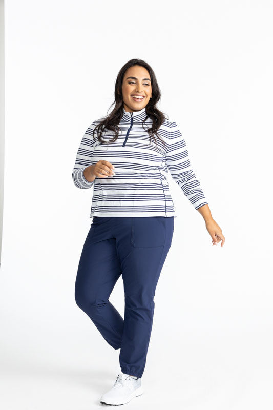 Smiling woman wearing the Keep It Covered Long Sleeve Golf Top in Shutter Stripe and the Tailored and Trip Jogger Pants in Navy Blue. This pattern is a grouping of four horizontal, navy blue stripes with white space in between from top to bottom on this t