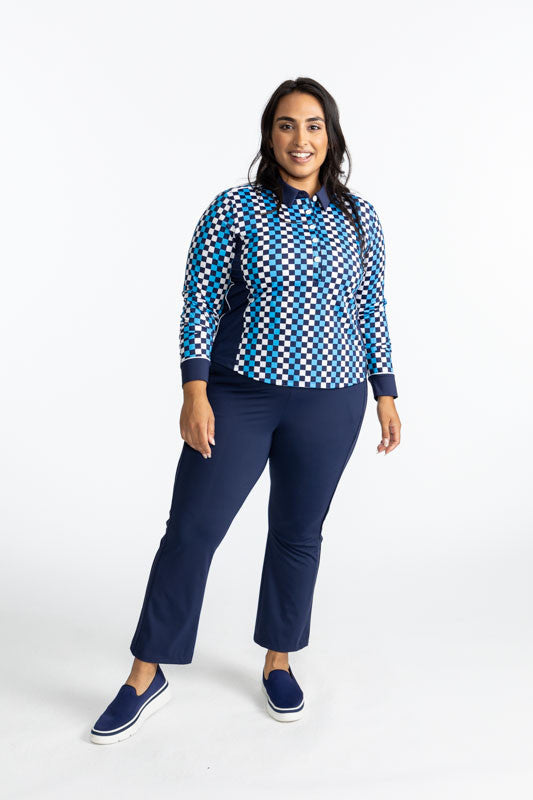 Full front view of a smiling woman wearing the Tailored Track Golf Pants in Navy Blue and the At The Pin Long Sleeve Golf Top in Check It Out print. This print is a mix of French blue, black, and white checks forming a vertical striped pattern. This shirt