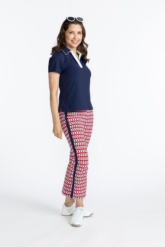 Smiling woman with sunglasses on her head wearing the Classic and Fantastic Short Sleeve Golf Top in Navy Blue and the Smooth Your Waist Crop Pants in Chevron Tomato Red. There are also white accents on the edge of the collar and the v-neck on this top.