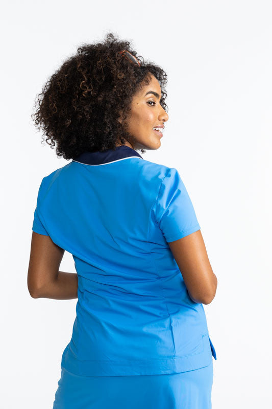 Back view of a woman wearing the the Classic and Fantastic Short Sleeve Golf Top in French Blue. This top is a solid French blue with a navy blue collar trimmed with white and a navy blue V-neck front.