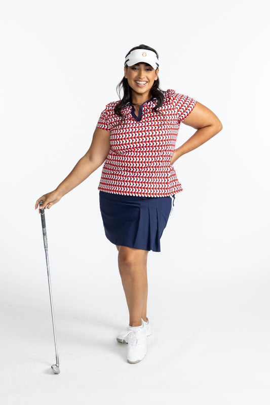 Full front view of a smiling woman golfer holding a club in her right hand wearing the Classic and Fantastic Short Sleeve Golf Top in Chevron Tomato Red, the Party Pleat Golf Skort in Navy Blue, and the No Hat Hair Visor in White. This top has a navy blue