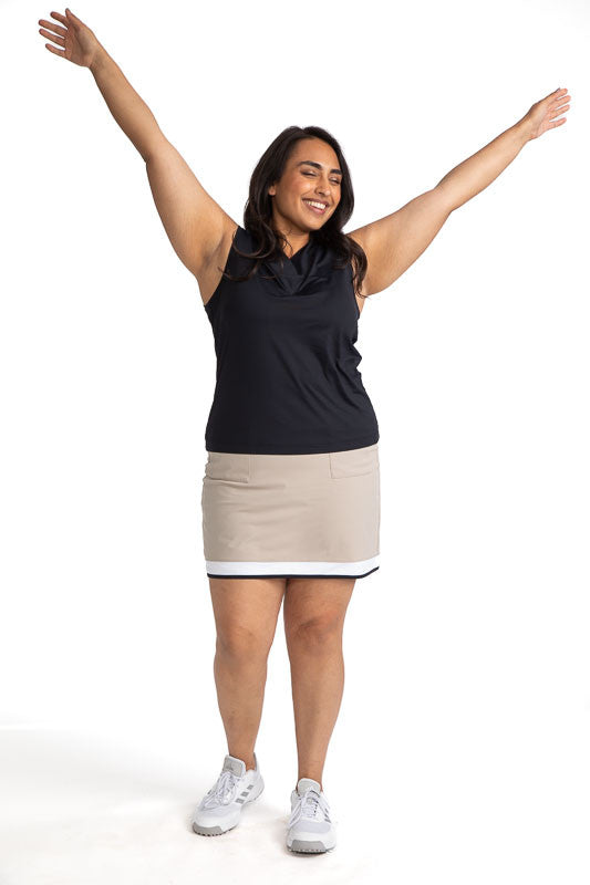 Smiling woman golfer with both arms up in the air wearing the Tee to Tea Golf Skort in Sand and the Light and Lovely Sleeveless Golf Top in Black.  This skort has white and black accents across the top of each front pocket, a large, white band, and a smal