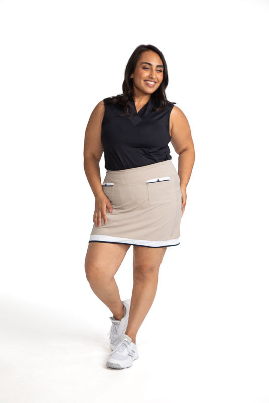 Smiling woman golfer wearing the Tee to Tea Golf Skort in Sand and the Light and Lovely Sleeveless Golf Top in Black.  This skort has white and black accents across the top of each front pocket, a large, white band, and a smaller, black band around the he