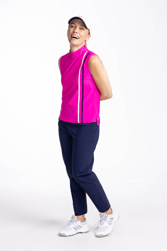 Full front and left side view of a smiling woman wearing the No Break Sleeveless Golf Top in Open Air Pink, the Tailored Crop Golf Pants in Navy Blue, and the We've Got You Covered Hat in White. This is a bright pink top with two vertical stripes in white