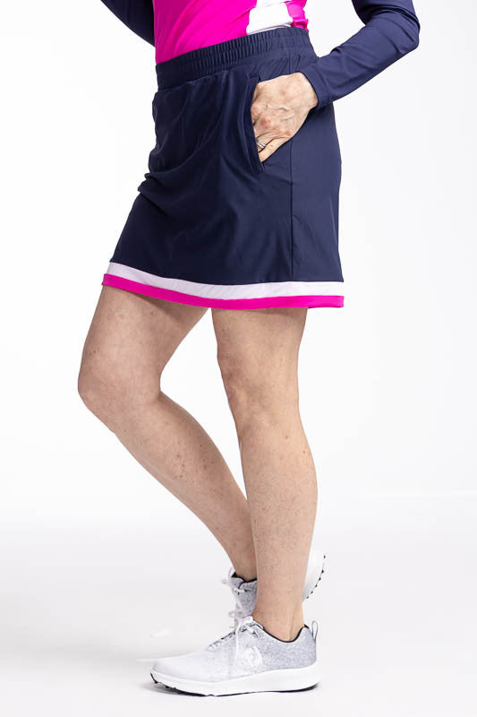 Left side view of the Comfy and Cozy Golf Skort in Navy Blue. This is a navy blue skort with two horizontal stripes around the hemline - one white and one open air pink. It also features two side zippered pockets with a built-in tee holder, two back zippe