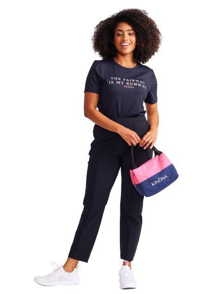 Full front view of a woman wearing The Fairway is my Runway t-shirt in black, the Tailored Crop Pants in black, and holding the Snack Sack in Open Air Pink in her hands on the left side of her body.