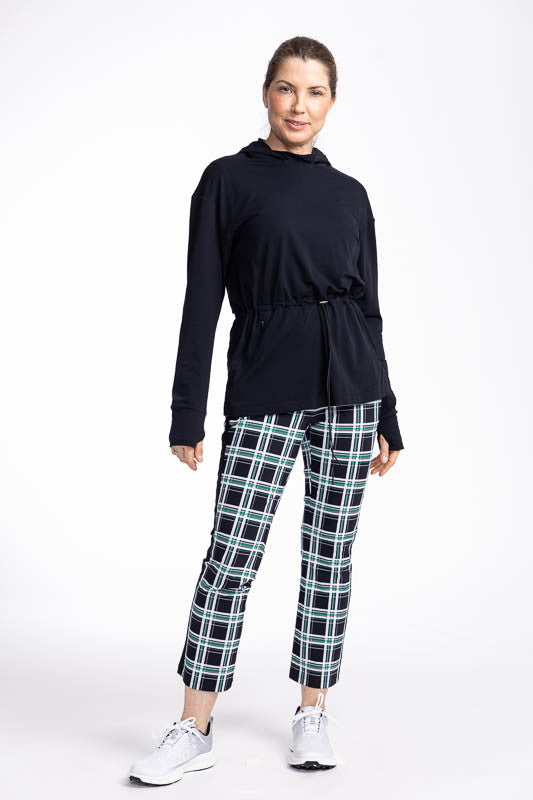 Full front view of a woman wearing the Aprés 18 Anorak Long Sleeve Hoodie in black and the Smooth Your Waist Crop Golf Pants in Tartan Plaid.