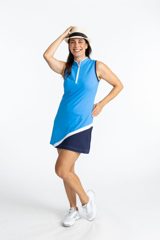 Full front view of a woman with her right hand on her straw hat and her left hand on her hip wearing the Tend The Flag Sleeveless Golf Dress in French Blue. This dress has a white quarter zip on the front of the dress, navy blue accents around each arm ho
