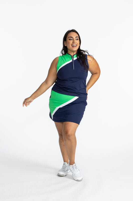 Smiling woman golfer wearing the Tend the Flag Golf Skort in Navy Blue and the Tend the Flag Sleeveless Golf Top in Navy Blue. This top features a navy blue background with a diagonal white stripe and a section of Kelly green from the top of the left shou