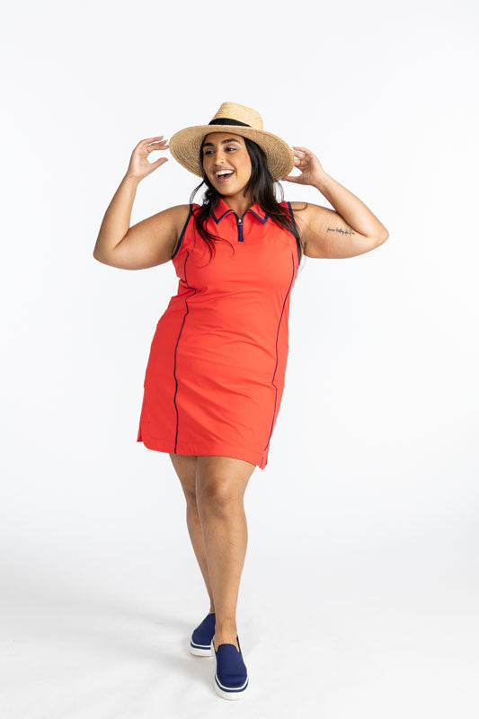 Full front view of a smiling woman wearing a straw hat and the In The Cup Sleeveless Golf Dress in Tomato Red. This dress has black accents around the collar and down the front quarter zip, around each armhole, and one black line down each side on the fro