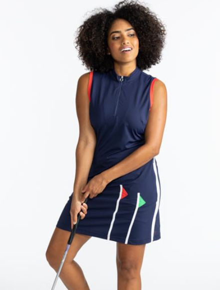 Full front view of a smiling woman wearing the Flagstick Sleeveless Golf Dress in Navy Blue. This dress has a navy blue background, tomato red around each arm hole, one white stripe that runs the full length of the dress on each side, and two golf flagsti