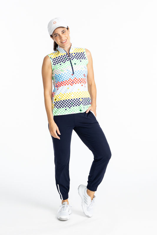 Smiling woman golfer wearing the We've Got You Covered Hat in White, the Tailored and Trim Jogger Golf Pants in Navy Blue, and the Keep It Covered Sleeveless Golf Top in Cheeky Check Print. This print is made up of a checked horizontal patter that creates