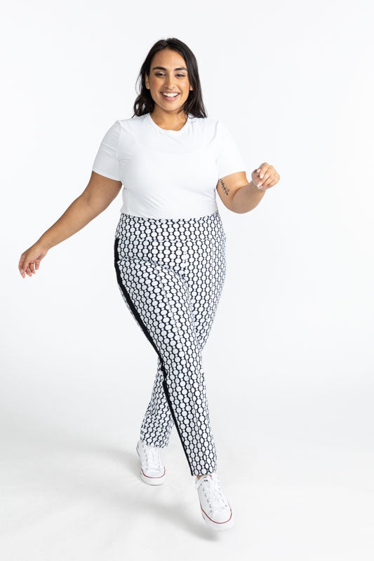 Smiling woman mid-stride wearing the Tee it Up Short Sleeve Golf Shirt in White and the Tailored Track Golf Pants in Spiral Floral. This print is a retro-inspired abstract black and white flower pattern. There is one black stripe that runs the length on e