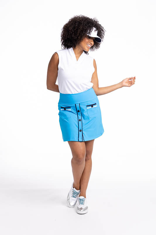 Full front view of a smiling woman wearing the Long Strides Golf Skort in Pacific Blue, the Light and Lovely Sleeveless Golf Top in White, and the No Hat Hair Visor in White. This skort is a solid blue with two patch pockets that have a navy blue stripe a