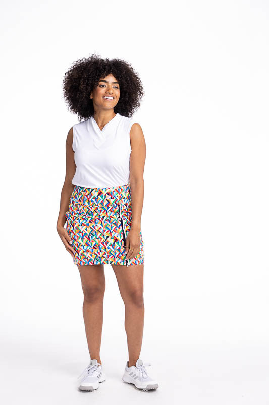 Full front view of a smiling woman wearing the Summer Sass Golf Skort in K All Day print and the Light and Lovely Sleeveless Golf Top in White. The K All Day print is an abstract confetti-style mirrored K-shaped pattern in red, orange, yellow, green, blue