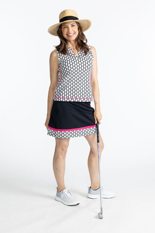 Full front view of a smiling woman holding a golf club in her left hand wearing a straw hat, the In Play Golf Skort in Black, and the Bogey Round Sleeveless Golf Top in Spiral Floral. This is a retro-inspired abstract floral pattern in black and white.