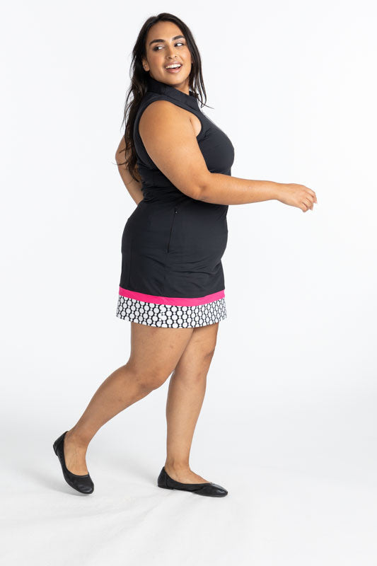 Full right side view of a woman wearing the In Play Sleeveless Golf Dress in Black. This dress is primarily black with a white 1/4 zip front, a thin band of preppy pink ear the bottom of the dress, followed by a thicker band of spiral floral (black and wh