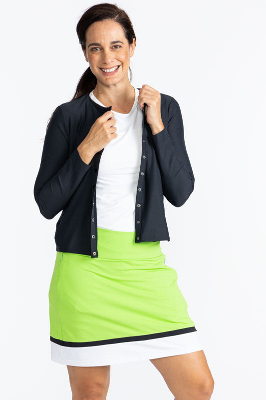 Full front view of a smiling woman wearing the Party  Cardi in Black, the Tee it Up Golf Shirt in White, and the In Play Golf Skort in Grass Green