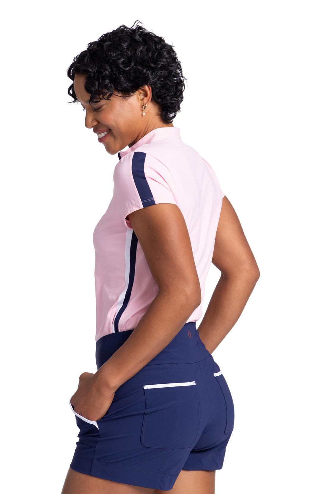 Side view of a woman wearing a soft pink, short sleeve top. The top has a navy blue stripe going down the middle of the sleeve and contrasting white and navy blue stripes going down the side. The top is worn with navy blue shorts with white trim on the back and side pockets. 