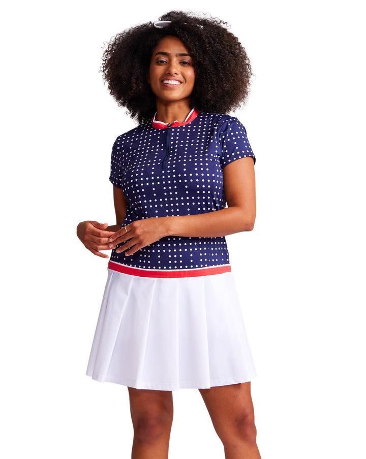 Front view of a woman wearing an above the knee, drop waist, domino navy with white dots dress with white pleating on the bottom and red trim on the neckline and waist. 