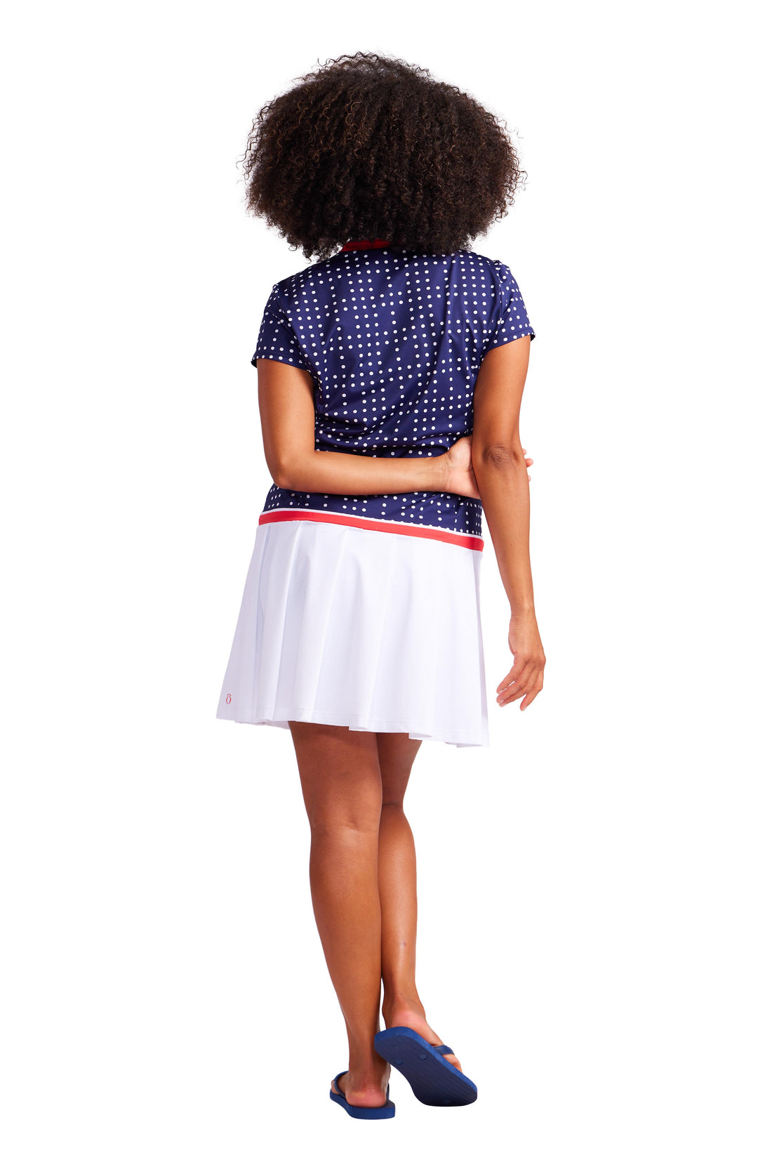 Back view of a woman wearing an above the knee, drop waist, domino navy with white dots dress with white pleating on the bottom and red trim on the neckline and waist.