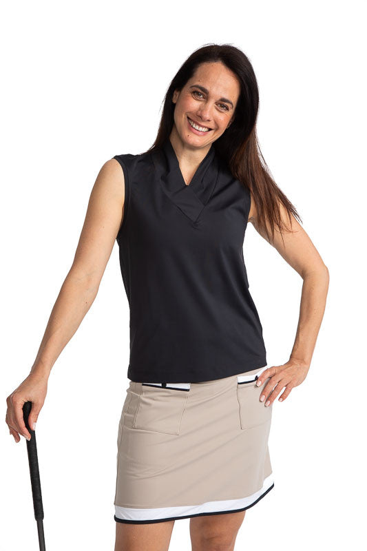 Front view of a smiling woman golfer holding a club in her right hand with her left hand on her hip wearing the Light and Lovely Sleeveless Golf Top in Black and the Tee to Green Golf Skort in Sand.